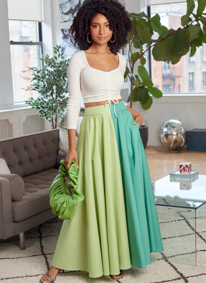 May include: A woman wearing a white long-sleeved crop top with a tie at the waist and a green and blue maxi skirt. She is holding a green handbag.