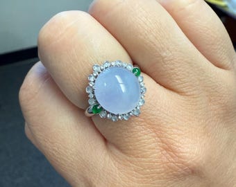 May include: A silver ring featuring a large, oval, pale blue gemstone surrounded by small, clear stones and two small green stones. The ring is set on a hand.