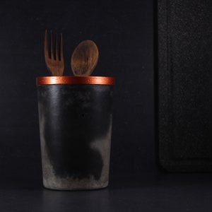 May include: A black utensil holder with a copper rim, holding a wooden spoon and fork.