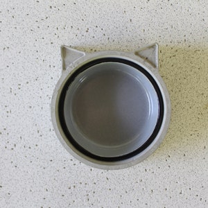 K&ouml;nnte beinhalten: Eine graue, katzenf&ouml;rmige Sch&uuml;ssel aus Keramik mit schwarzem Rand. Die Sch&uuml;ssel ist leer und steht auf einer wei&szlig;en, gefleckten Oberfl&auml;che.