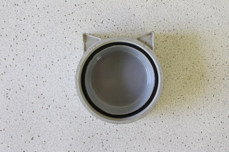 K&ouml;nnte beinhalten: Eine graue, katzenf&ouml;rmige Sch&uuml;ssel aus Keramik mit schwarzem Rand. Die Sch&uuml;ssel ist leer und steht auf einer wei&szlig;en, gefleckten Oberfl&auml;che.
