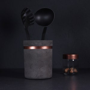 May include: A gray concrete utensil holder with a copper band around the middle. The holder has a black ladle and a black pasta server inside. A small glass jar with a copper lid is to the right of the holder.