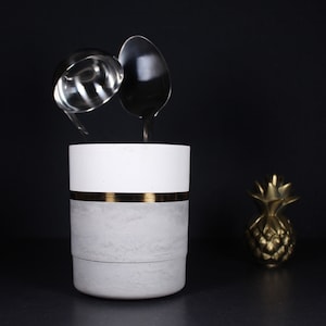 May include: A modern kitchen utensil holder with a white top, gray base, and gold band. Two silver spoons are suspended above the holder. A gold pineapple figurine is to the right against a black background.