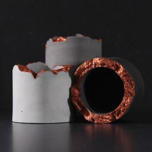 以下が含まれることがあります： 銅色の縁取りが施されたコンクリート製のプランターが3つ。プランターはサイズが異なり、積み重ねられています。