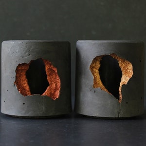 May include: Two grey concrete cylindrical planters with irregular holes. The holes are lined with copper and gold metallic paint.