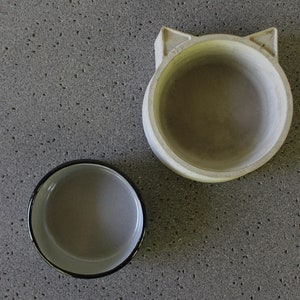 Exploded view of a two-piece pet feeding set featuring a minimalist light grey concrete base with a unique cat-ear silhouette and a separate grey enamel metal bowl insert, displayed side-by-side on a textured stone surface.