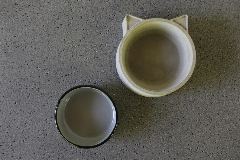 Exploded view of a two-piece pet feeding set featuring a minimalist light grey concrete base with a unique cat-ear silhouette and a separate grey enamel metal bowl insert, displayed side-by-side on a textured stone surface.