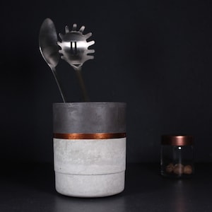 Könnte beinhalten: Ein grauer Beton-Utensilienhalter mit einem Kupferband um die Mitte. Der Halter enthält zwei Metallutensilien, einen Löffel und einen Schöpflöffel.