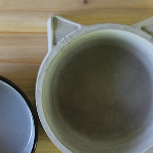 ​A top-down exploded view of the two-piece pet feeder set on a light wood surface, showing the grey enamel bowl insert placed next to the handcrafted concrete base with its distinct cat-ear silhouette. This shot clearly displays the interior of both the non-porous enamel bowl and the smooth, solid concrete holder.