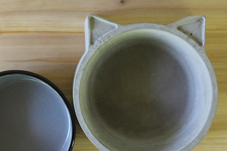 ​A top-down exploded view of the two-piece pet feeder set on a light wood surface, showing the grey enamel bowl insert placed next to the handcrafted concrete base with its distinct cat-ear silhouette. This shot clearly displays the interior of both the non-porous enamel bowl and the smooth, solid concrete holder.