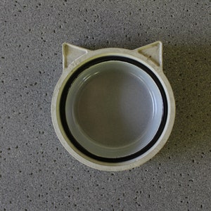 K&ouml;nnte beinhalten: Wei&szlig;e Keramik Sch&uuml;ssel in Katzenform mit schwarzem Rand. Die Sch&uuml;ssel ist leer und hat eine glatte, gl&auml;nzende Oberfl&auml;che.