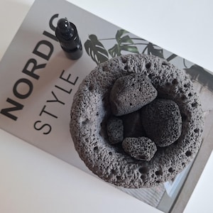 Puede incluir: Un difusor de cerámica negro con una superficie redonda y texturizada. El difusor contiene varias piedras de lava pequeñas y negras. También se puede ver una pequeña botella negra con un gotero en la imagen.