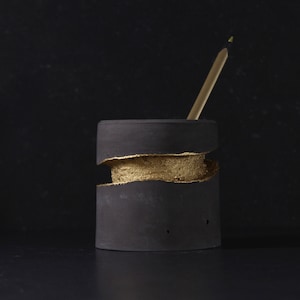 May include: A black and gold pencil holder with a crack running around the middle. The holder is made of concrete and has a gold accent.