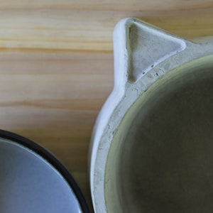 Close-up macro shot of the handcrafted concrete pet feeder, focusing on the textured triangular cat-ear detail and the smooth rim of the base. The image highlights the organic, stone-like texture of the polymer-enhanced concrete and its artisanal finish against a soft wooden background.