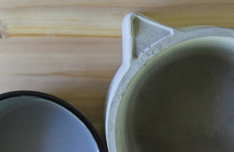 Close-up macro shot of the handcrafted concrete pet feeder, focusing on the textured triangular cat-ear detail and the smooth rim of the base. The image highlights the organic, stone-like texture of the polymer-enhanced concrete and its artisanal finish against a soft wooden background.