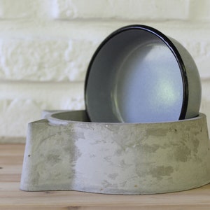 Side profile of a handcrafted grey concrete pet feeder showing the depth of the cat-shaped base and the removable grey enamel bowl insert, highlighting the raw and organic texture of the material.