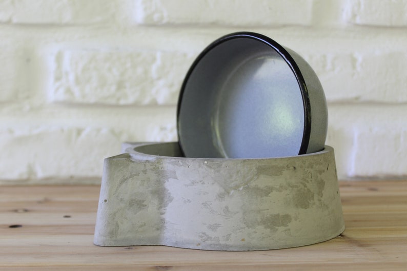 Side profile of a handcrafted grey concrete pet feeder showing the depth of the cat-shaped base and the removable grey enamel bowl insert, highlighting the raw and organic texture of the material.