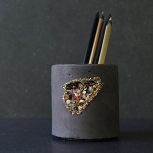Puede incluir: Un porta lápices de hormigón gris con una abertura con borde dorado llena de piedras de colores. El porta lápices está sobre una superficie negra.