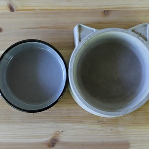 ​A top-down exploded view of the two-piece pet feeder set on a light wood surface, showing the grey enamel bowl insert placed next to the handcrafted concrete base with its distinct cat-ear silhouette. This shot clearly displays the interior of both the non-porous enamel bowl and the smooth, solid concrete holder.