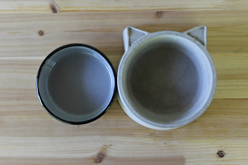 ​A top-down exploded view of the two-piece pet feeder set on a light wood surface, showing the grey enamel bowl insert placed next to the handcrafted concrete base with its distinct cat-ear silhouette. This shot clearly displays the interior of both the non-porous enamel bowl and the smooth, solid concrete holder.