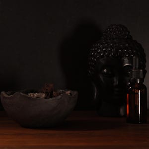 Op de afbeelding: Een donker stilleven met een zwart Boeddha-hoofd, een kleine bruine glazen fles met een druppelaar en een donkergrijze kom gevuld met gedroogde botanische producten. De objecten staan op een houten oppervlak.