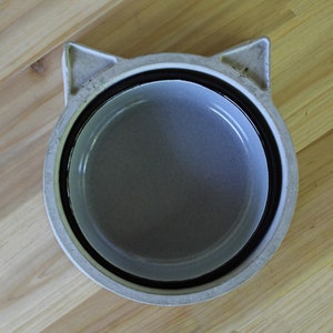 Top-down view of a handcrafted minimalist concrete pet bowl in light grey, featuring a unique cat-shaped design with subtle ear details. The bowl includes a removable grey enamel insert and is placed centrally on a rustic light wood plank surface, highlighting the contrast between the industrial concrete and natural wood.