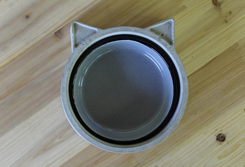 Top-down view of a handcrafted minimalist concrete pet bowl in light grey, featuring a unique cat-shaped design with subtle ear details. The bowl includes a removable grey enamel insert and is placed centrally on a rustic light wood plank surface, highlighting the contrast between the industrial concrete and natural wood.
