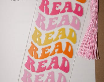 Peut inclure: Marque-page transparent avec le mot "READ" en police rétro, répété en orange, rose et jaune. Une pompon rose est attachée en haut. Le marque-page est placé sur une page de texte.