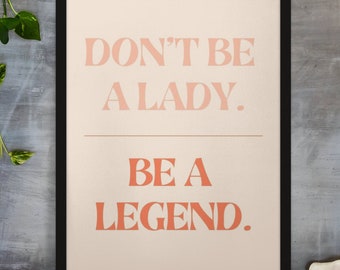 May include: A framed print with the text "DON'T BE A LADY. BE A LEGEND." in coral-colored letters on a cream background. The black frame sits on a wooden surface, with greenery in the background.