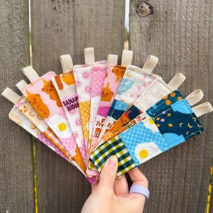 May include: A collection of colorful fabric coasters with various patterns, including teddy bears, fried eggs, and text that reads "BANAN BUDDI". Each coaster has a loop for hanging. The coasters are held in a hand.
