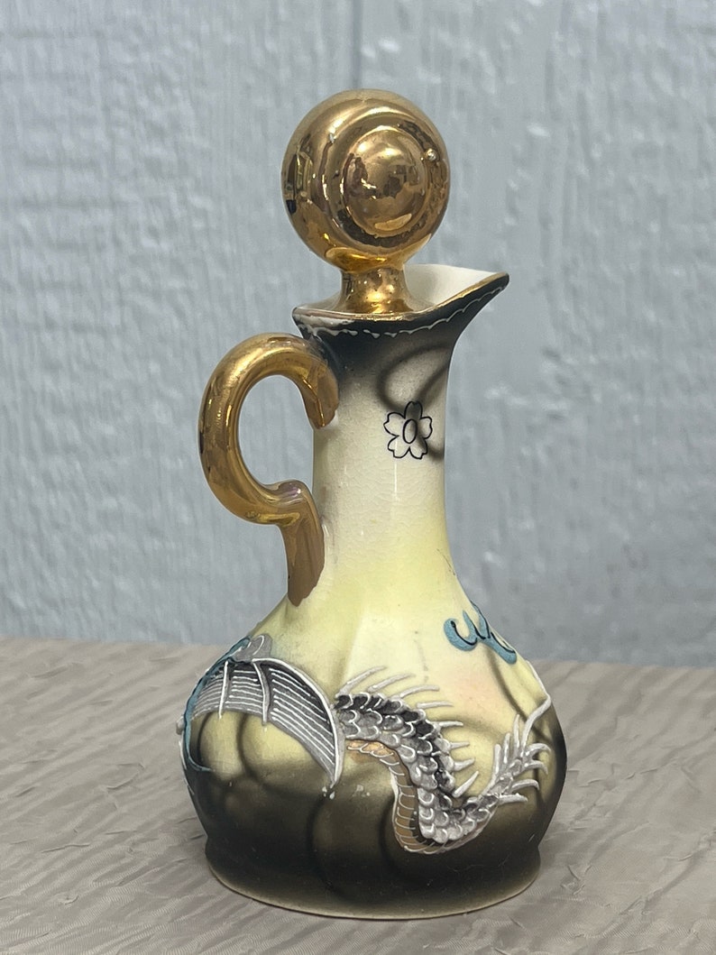 May include: A small, yellow ceramic bottle with a gold lid and a dragon design. The bottle has a gold handle and a floral design on the neck.