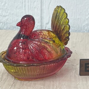 K&ouml;nnte beinhalten: Eine dekorative Glasschale in Form eines Truthahns mit Deckel. Die Schale ist haupts&auml;chlich rot und gelb, mit einem detaillierten Federdesign. Der Boden hat eine Schalenform, und daneben befindet sich ein kleiner Holzblock mit dem Buchstaben "B".