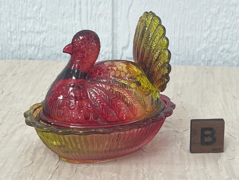 K&ouml;nnte beinhalten: Eine dekorative Glasschale in Form eines Truthahns mit Deckel. Die Schale ist haupts&auml;chlich rot und gelb, mit einem detaillierten Federdesign. Der Boden hat eine Schalenform, und daneben befindet sich ein kleiner Holzblock mit dem Buchstaben "B".