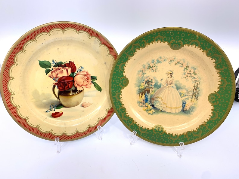 May include: Two vintage tin plates with decorative borders. One plate features a bouquet of red and pink roses in a gold vase. The other plate features a woman in a white dress standing in a garden setting.