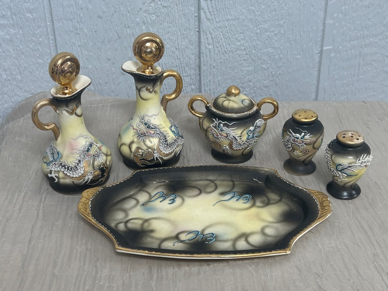 May include: A set of six porcelain dragon-themed serving pieces, including two small pitchers, a sugar bowl, two salt shakers, and a serving tray. The pieces are decorated with gold accents and a black and white dragon design.