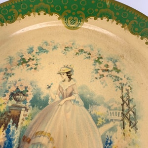 May include: A vintage tin tray with a green and gold border. The center of the tray features a painted image of a woman in a white hat and a light blue and pink dress standing in a garden setting.
