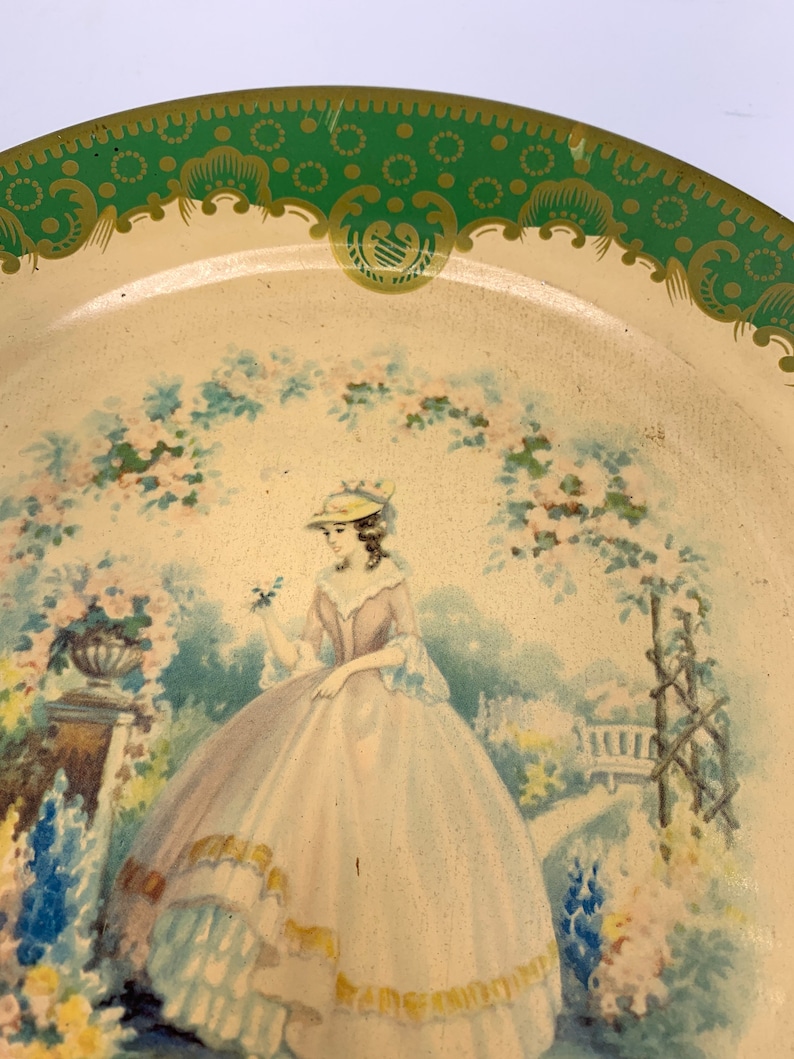 May include: A vintage tin tray with a green and gold border. The center of the tray features a painted image of a woman in a white hat and a light blue and pink dress standing in a garden setting.