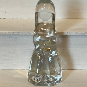 May include: A clear glass figurine of a woman with long hair and a flowing dress. The figurine is standing on a white surface.