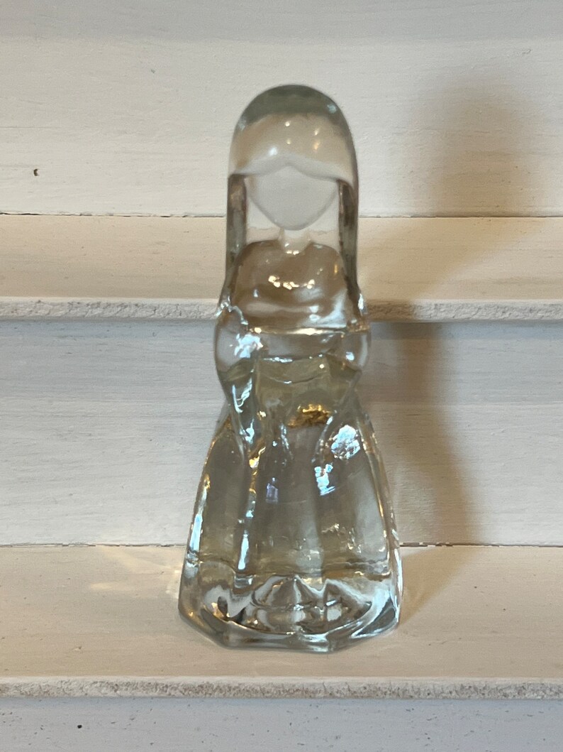 May include: A clear glass figurine of a woman with long hair and a flowing dress. The figurine is standing on a white surface.