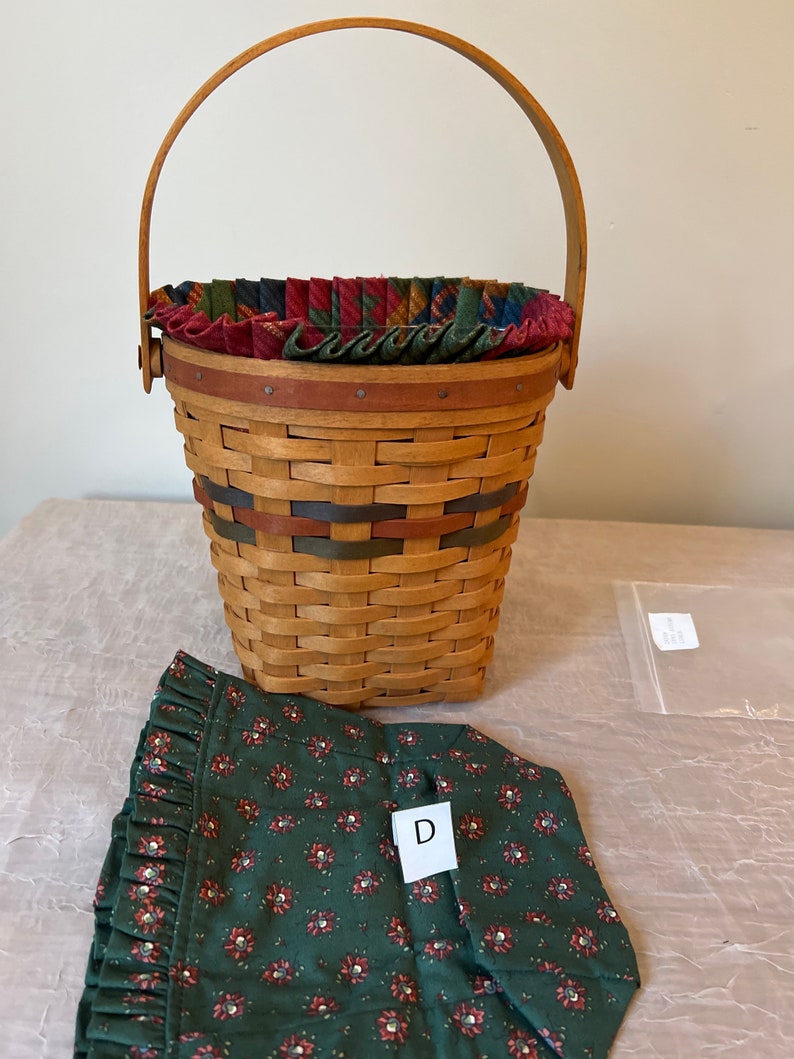 May include: A woven basket with a wooden handle and a fabric liner. The basket is brown and has a green and red patterned fabric liner. The basket is sitting on a table with a green fabric with red flowers on it. The fabric has a white tag with the letter "D" on it.