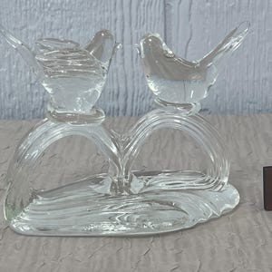 K&ouml;nnte beinhalten: Eine klare Glasskulptur mit zwei V&ouml;geln, die auf einer herzf&ouml;rmigen Basis sitzen. Die V&ouml;gel haben detaillierte Fl&uuml;gel und sind durch ein geschwungenes Design verbunden. Ein kleiner, brauner W&uuml;rfel mit dem Buchstaben "C" befindet sich neben der Skulptur.