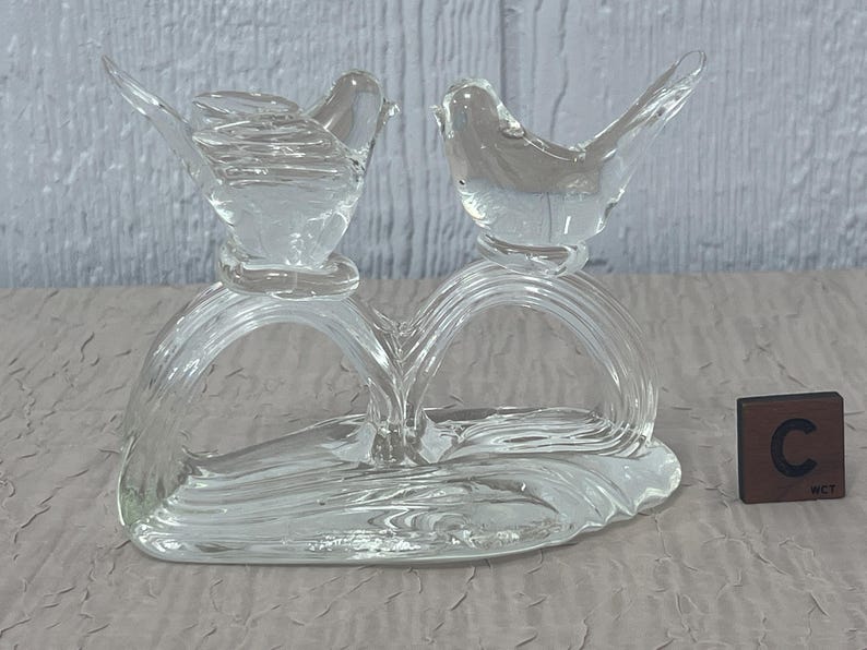 K&ouml;nnte beinhalten: Eine klare Glasskulptur mit zwei V&ouml;geln, die auf einer herzf&ouml;rmigen Basis sitzen. Die V&ouml;gel haben detaillierte Fl&uuml;gel und sind durch ein geschwungenes Design verbunden. Ein kleiner, brauner W&uuml;rfel mit dem Buchstaben "C" befindet sich neben der Skulptur.