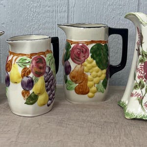 May include: Four ceramic pitchers with fruit designs. Three pitchers have black handles and are decorated with grapes, pears, and apples. The fourth pitcher is white with a red rose design and a green handle.