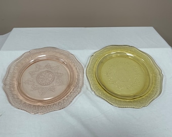 May include: Two vintage glass plates with a decorative pattern. The plate on the left is pink and the plate on the right is yellow.