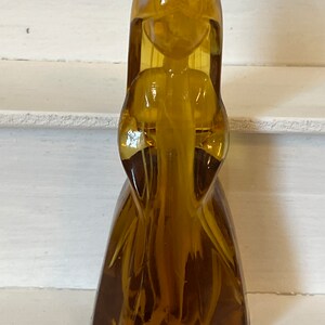 Pu&ograve; includere: Una statuetta vintage in vetro color ambra a forma di figura stilizzata con capelli lunghi e un abito fluente. La statuetta ha una finitura liscia e lucida e si erge in posizione verticale. I lineamenti della figura sono semplici ed eleganti.