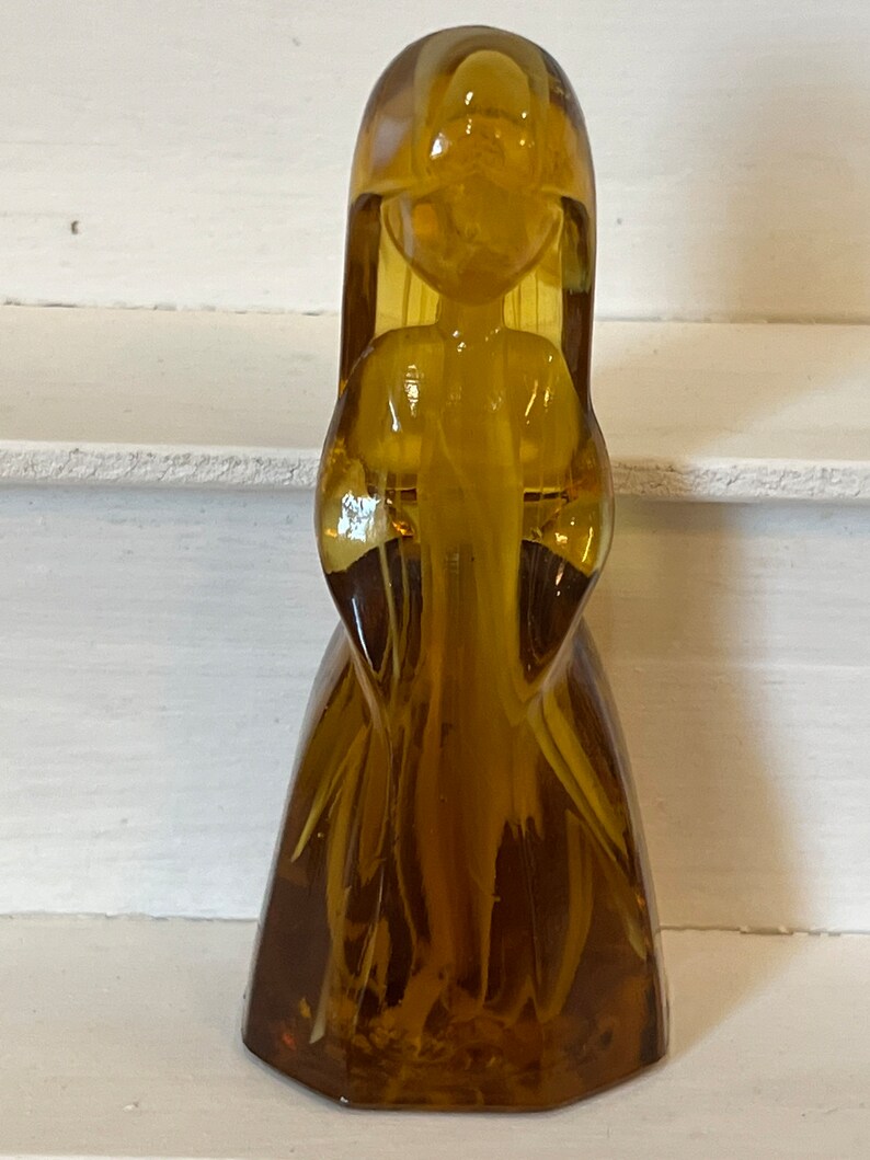 Pu&ograve; includere: Una statuetta vintage in vetro color ambra a forma di figura stilizzata con capelli lunghi e un abito fluente. La statuetta ha una finitura liscia e lucida e si erge in posizione verticale. I lineamenti della figura sono semplici ed eleganti.
