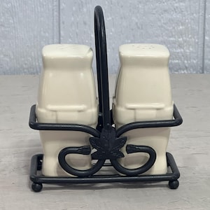 May include: A pair of off-white ceramic salt and pepper shakers held in a black metal caddy. The caddy features a central handle with a decorative leaf-shaped accent at the base and a wire frame that secures each shaker in an upright position.