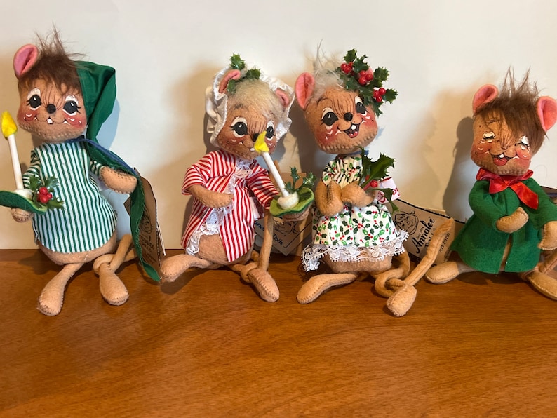 K&ouml;nnte beinhalten: Vier Pl&uuml;schmaus-Figuren, die in festliche Weihnachtskleidung gekleidet sind. Die M&auml;use tragen gr&uuml;n-rot-wei&szlig; gestreifte Pyjamas, ein gr&uuml;nes Kleid mit Stechpalmen-Akzenten, ein rot-wei&szlig; gestreiftes Kleid mit wei&szlig;er Spitzenborte und einen gr&uuml;nen Mantel mit einer roten Schleife. Die M&auml;use halten Kerzen, Stechpalmenzweige und ein kleines wei&szlig;es Geschenk.