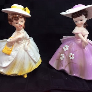 May include: Two vintage ceramic planter figurines of women wearing hats and dresses. One figurine is wearing a yellow dress and a white hat with a yellow ribbon. The other figurine is wearing a purple dress with white flowers and a white hat with a purple ribbon.