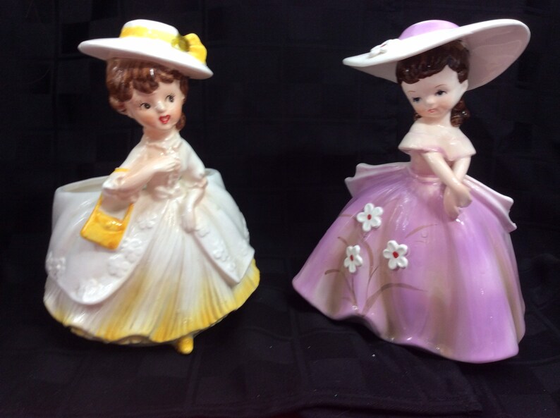 May include: Two vintage ceramic planter figurines of women wearing hats and dresses. One figurine is wearing a yellow dress and a white hat with a yellow ribbon. The other figurine is wearing a purple dress with white flowers and a white hat with a purple ribbon.